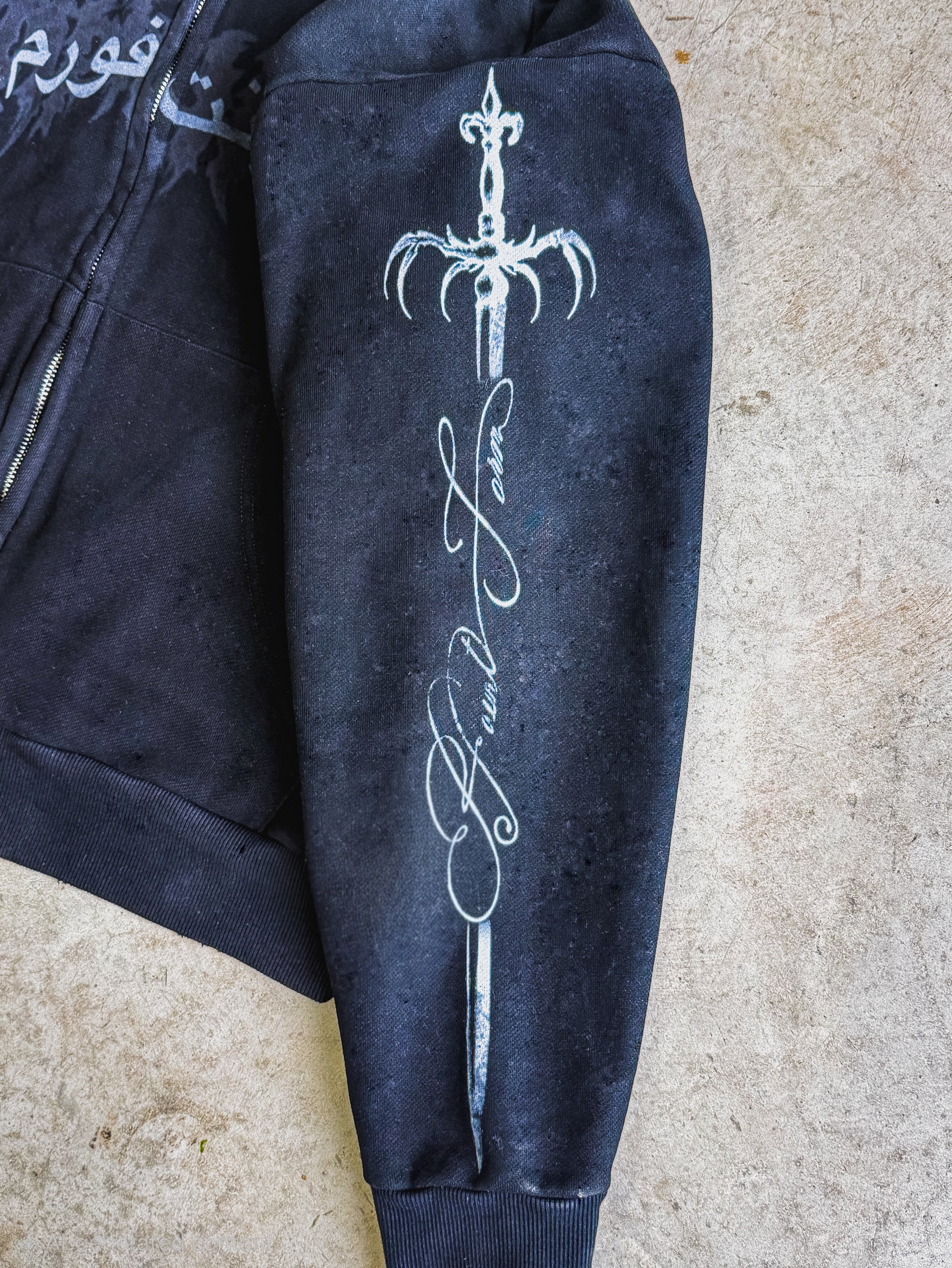 A close-up of a black zip-up hoodie laid flat on a textured beige surface, showing a large white graphic of a sword with Arabic calligraphy on the sleeve.